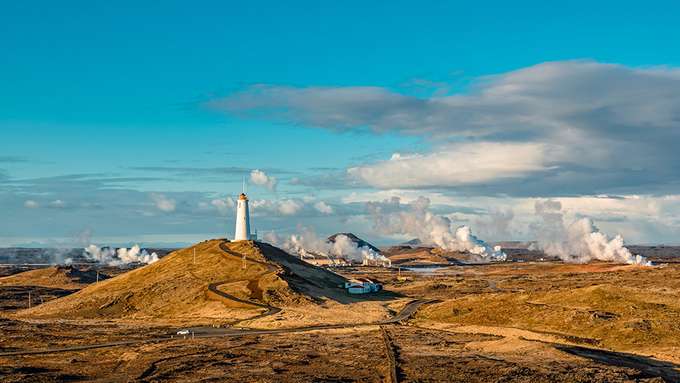 Landschaftbild mit weißem Leuchtturm, im Hintergrund Wasserdampf